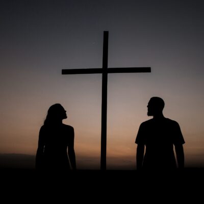 silhouette of man and woman standing beside cross during sunset